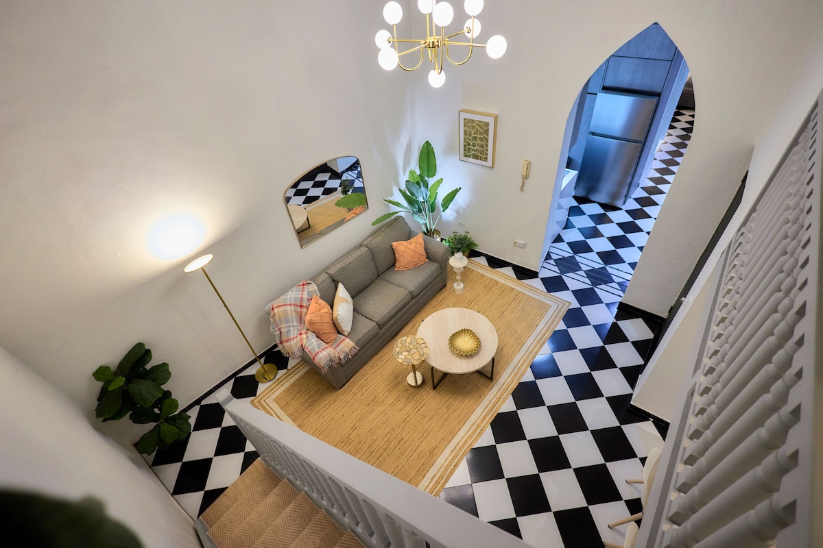An open and airy living space features a modern gray sofa adorned with colorful cushions. A round coffee table sits in the center, surrounded by a woven rug. A tall plant and floor lamp add warmth, while the black-and-white tiled floor creates a striking contrast.