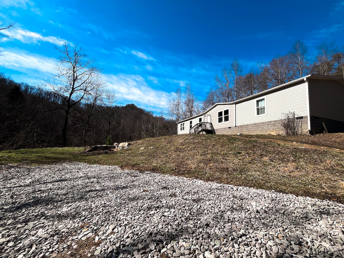 A modest cabin is featured against a backdrop of rolling hills and a vibrant blue sky, with scattered clouds. A gravel area leads up to the cabin, which is surrounded by bare trees and grassy patches, offering a sense of seclusion in nature.