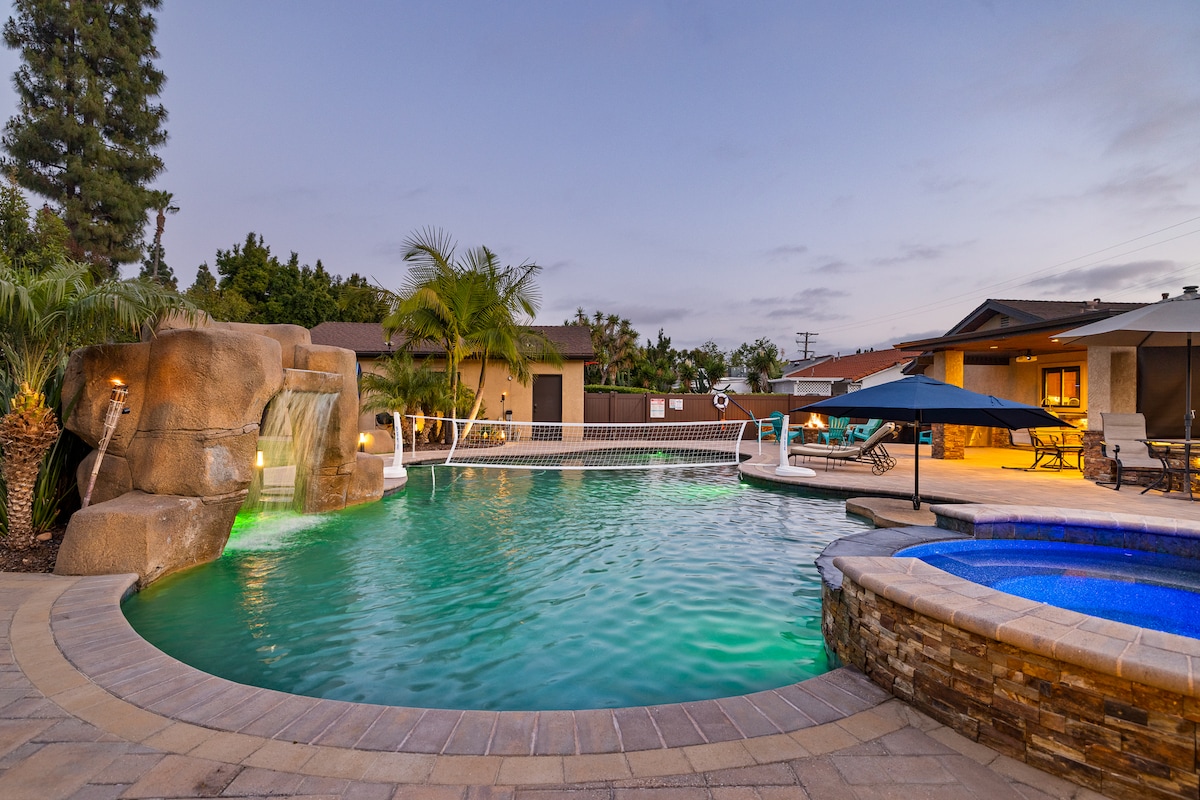 An inviting outdoor space showcases a large swimming pool featuring a waterfall and surrounding palm trees. A jacuzzi is positioned nearby, while comfortable lounge chairs are arranged under shaded umbrellas. Soft evening light enhances the tranquil atmosphere of the property.