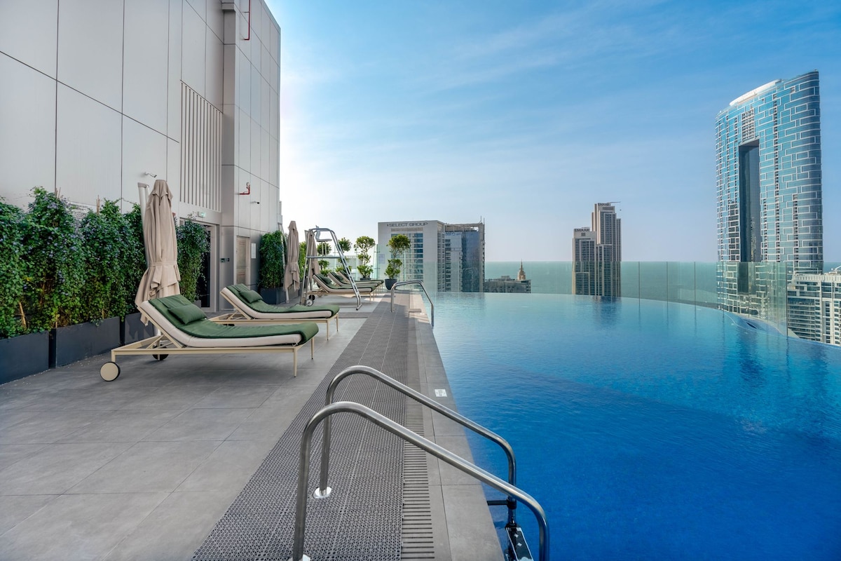 An infinity pool is displayed against a backdrop of the Dubai skyline, featuring sleek modern buildings. Lounge chairs line the edge, inviting relaxation. Lush greenery is placed along the pool area, enhancing the tranquil environment under a clear blue sky.