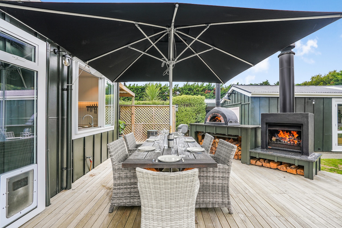 An outdoor deck area features a large table set for dining, accompanied by multiple wicker chairs. A large umbrella provides shade, while a stone fireplace and outdoor pizza oven are visible in the background, surrounded by greenery.