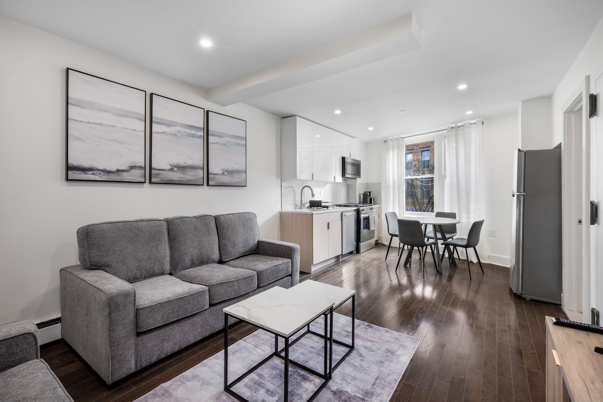 A modern living area is furnished with a gray sectional sofa and a marble-topped coffee table, arranged on a soft area rug. Natural light filters through sheer curtains, illuminating a dining table with chairs. An open kitchen is equipped with stainless steel appliances and sleek cabinetry.