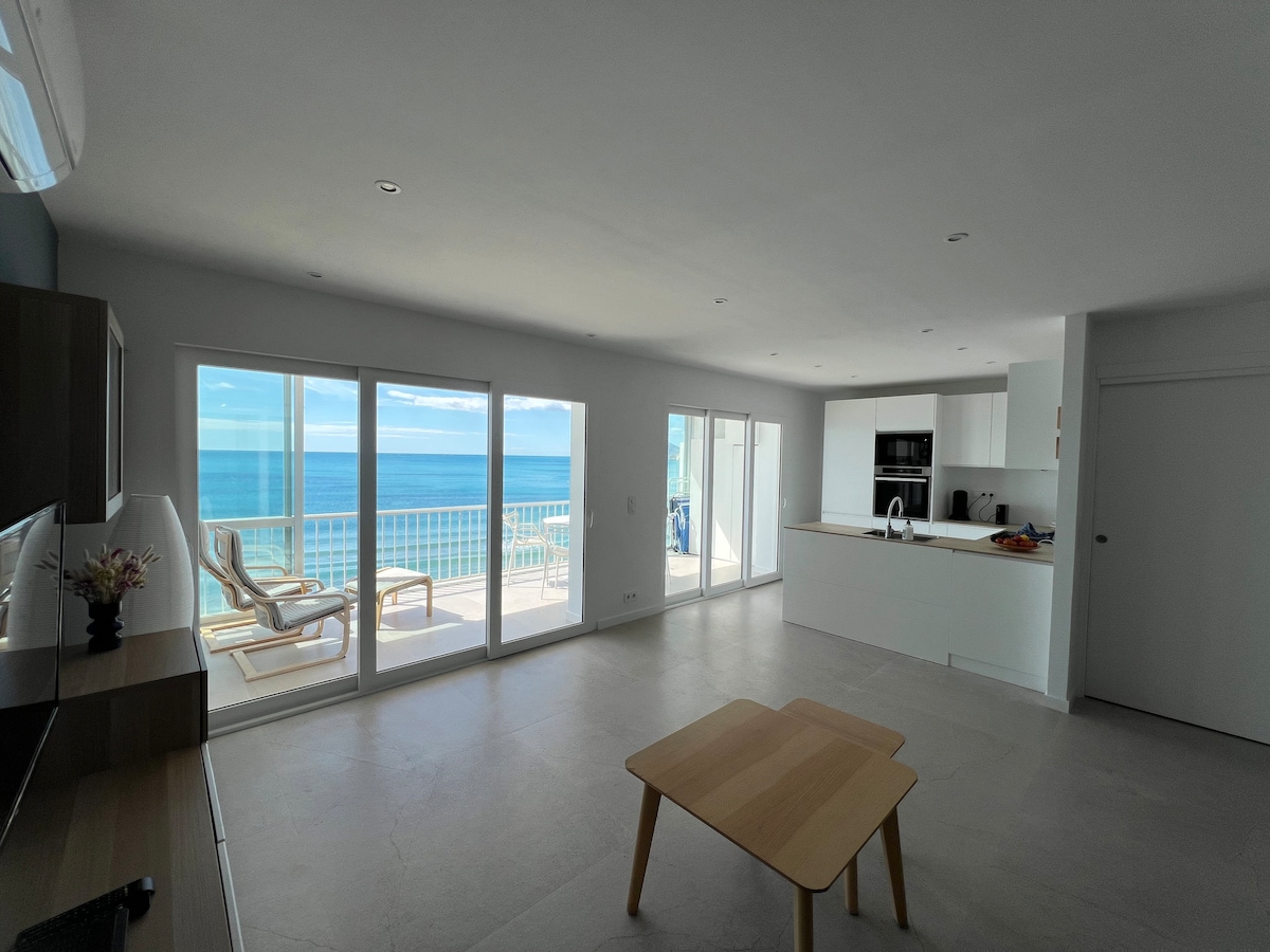 An open living area features large glass doors offering unobstructed views of the sea. The modern kitchen is equipped with sleek cabinetry and appliances, while a light wood table invites dining or relaxation. The space is bright and airy, with ample natural light flooding in.