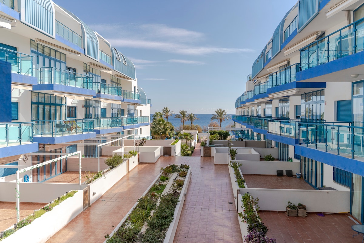 A scenic view of a modern apartment complex, featuring multiple levels with blue and white facades. Balconies with glass railings offer glimpses of the Mediterranean Sea, while landscaped areas with plants line the pathways, providing a serene atmosphere for residents and visitors.