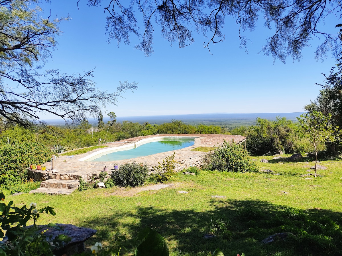 An outdoor oasis is depicted, featuring a spacious swimming pool framed by natural stone and lush greenery. The tranquil landscape extends in the background, showcasing expansive views of the surrounding valleys and hills under a clear blue sky.