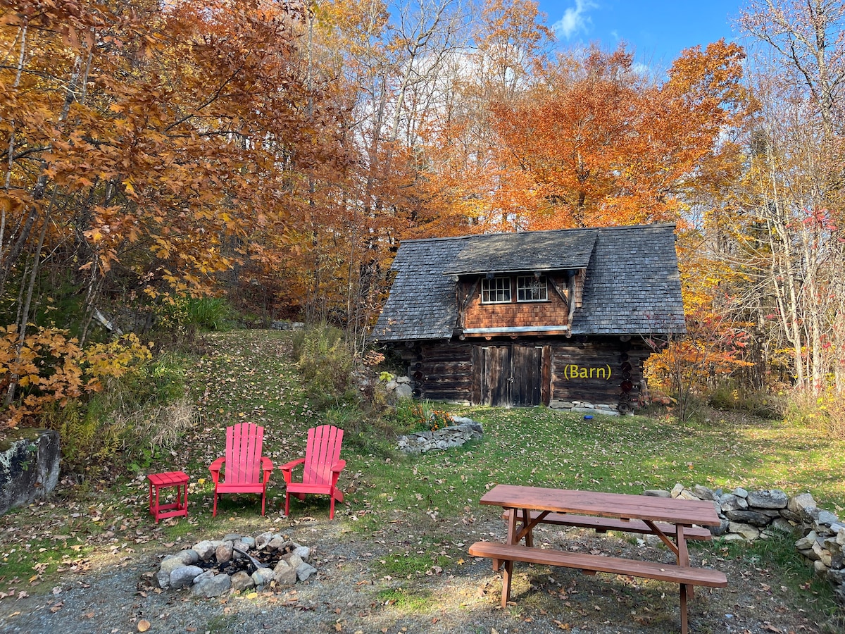 A rustic barn is nestled among vibrant autumn foliage. Nearby, two red Adirondack chairs surround a fire pit, with a picnic table positioned in front. The natural landscape captures the essence of the season, creating a serene outdoor gathering space.