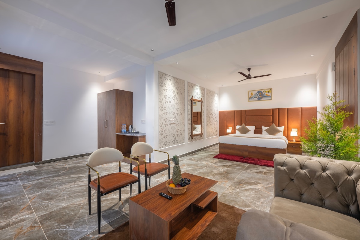 A spacious room features a large bed with soft bedding and ambient lighting. An elegant seating area includes a plush sofa and two chairs around a wooden coffee table. A stylish mirror accentuates the wall, while greenery adds a touch of nature to the atmosphere.