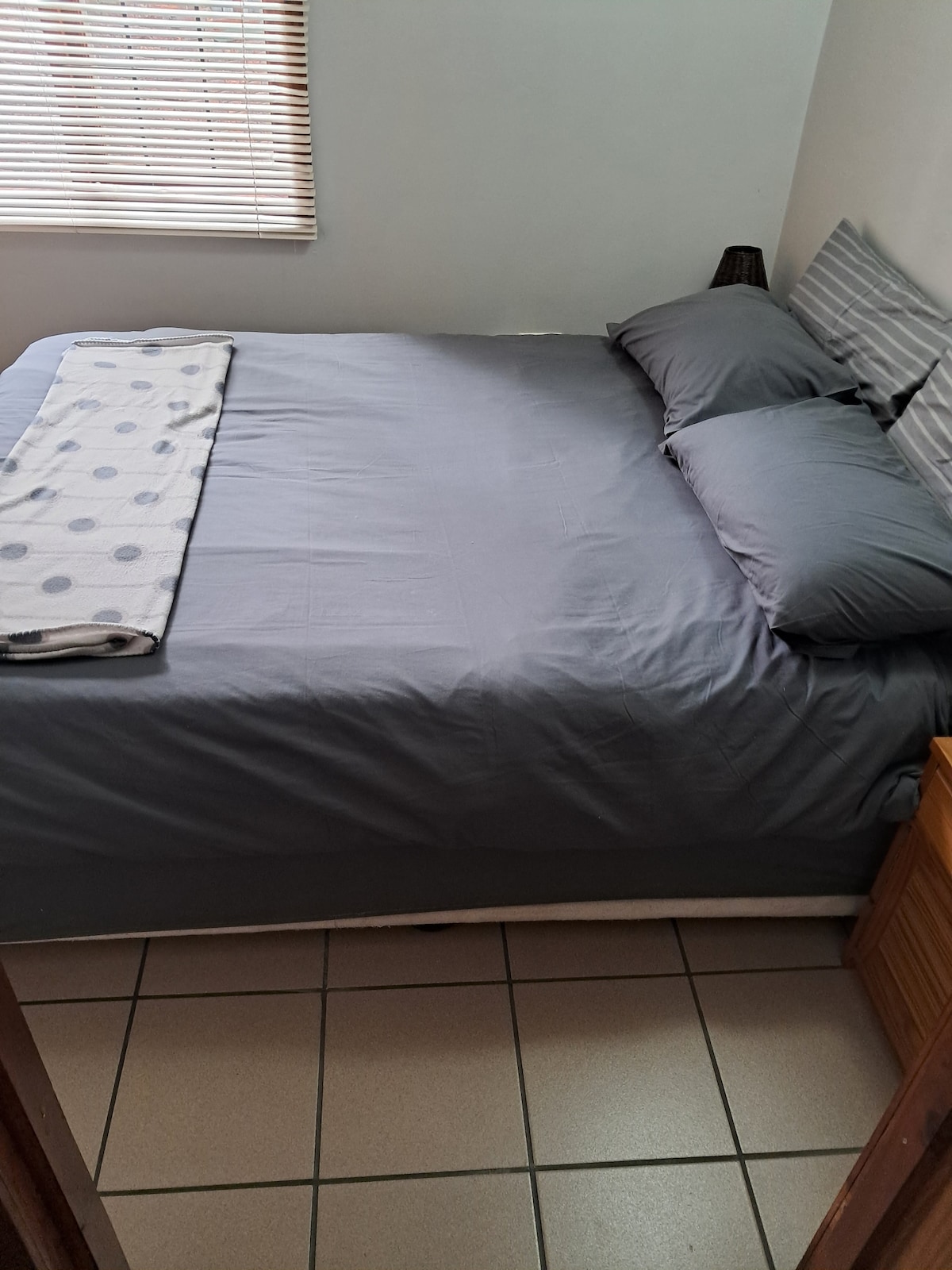 A cozy sleeping area features a spacious bed dressed in smooth gray linens, complemented by two matching pillows. Natural light filters through a window with wooden blinds, while a textured throw blanket is casually placed at the foot of the bed.