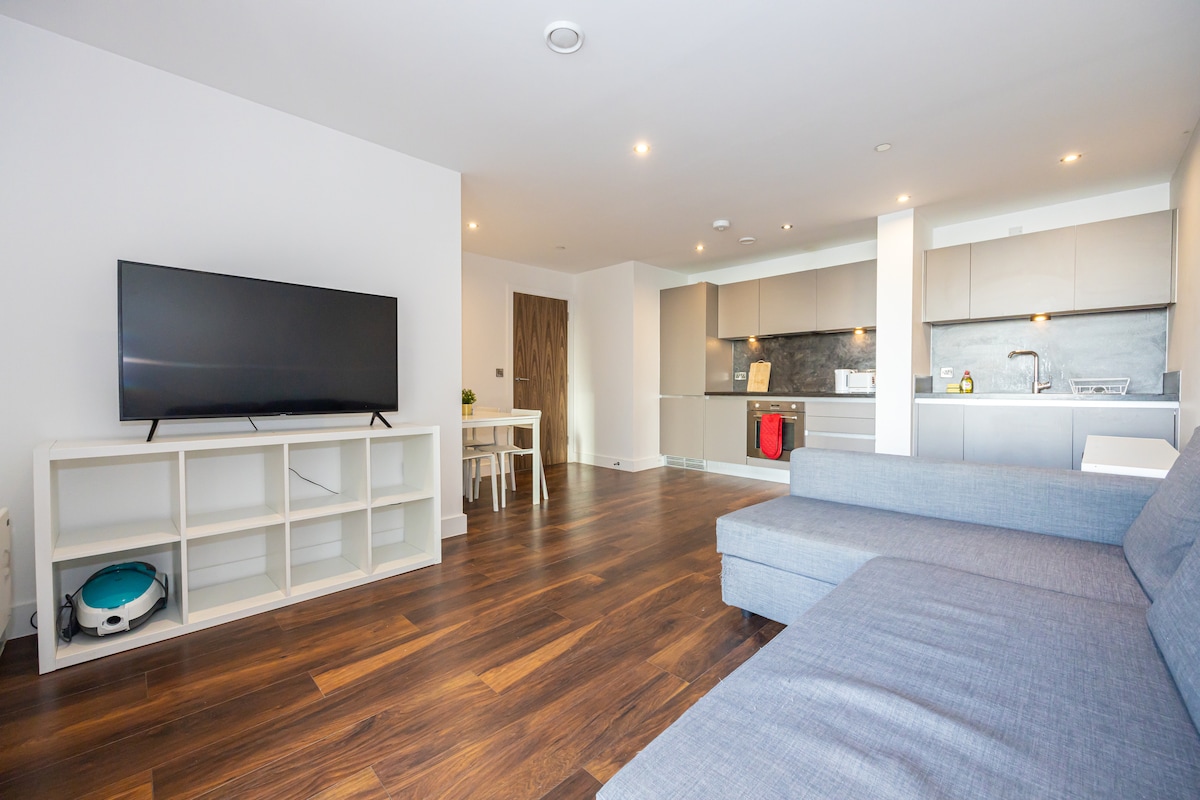 A spacious living area features a large flat-screen television on a white shelf. A modern kitchen is visible, equipped with essential appliances and a red dishwasher. A light-colored dining table with chairs is present, alongside a comfortable sectional sofa in a soft shade.