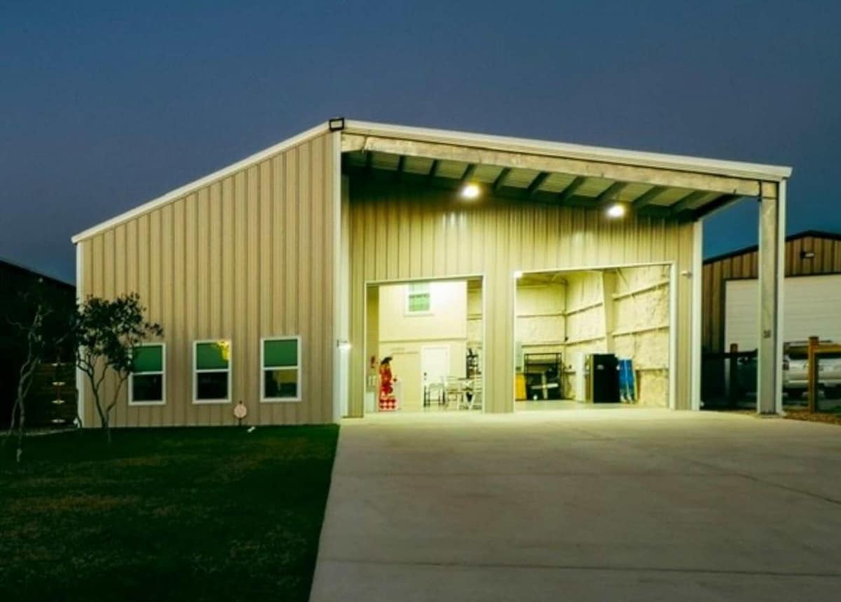 A spacious metal structure is set against a twilight sky. The open garage entrance reveals an organized interior with equipment and storage. The driveway leads up to the building, framed by green grass and illuminated by overhead lights.