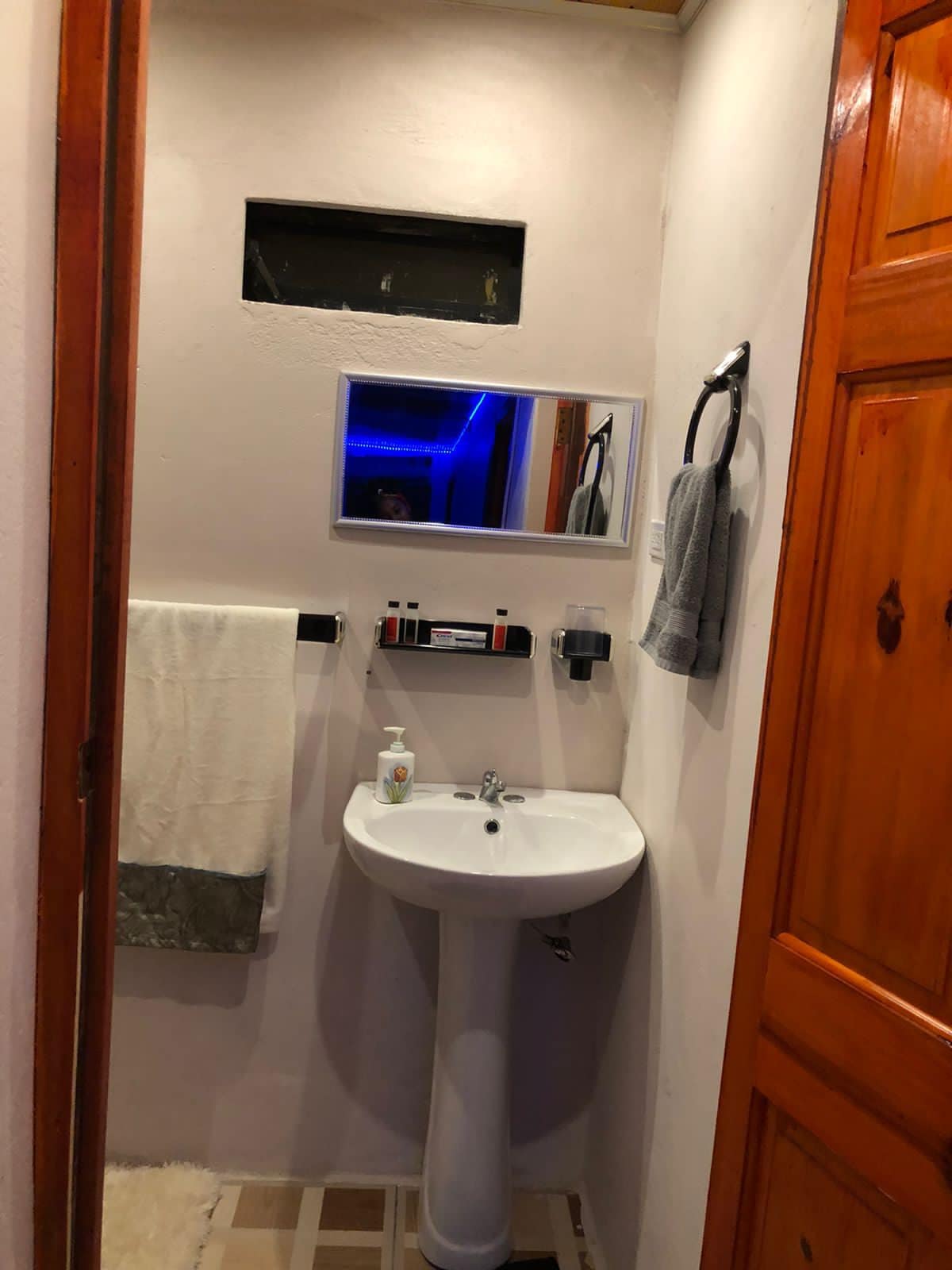 A compact bathroom is fitted with a white sink and a vanity mirror mounted on the wall. A hand towel is neatly hung beside the sink, and a soap dispenser is visible. Light blue ambient lighting is present above, with wooden cabinetry providing storage space.