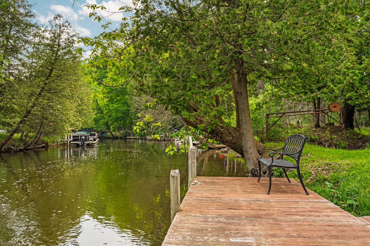 A wooden dock extends over calm waters, surrounded by lush green trees. A single black bench is positioned at the edge of the dock, providing a serene view of the nearby boats and the gentle flow of the river.