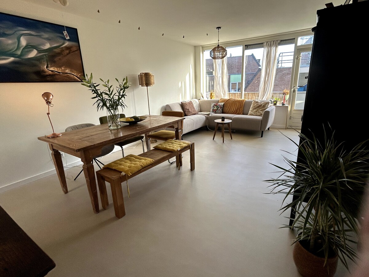 A bright living area is showcased, featuring a comfortable corner sofa adjacent to a dining table with wooden chairs. Natural light filters in through large windows, illuminating the neutral decor. A potted plant adds a touch of greenery, enhancing the inviting space.