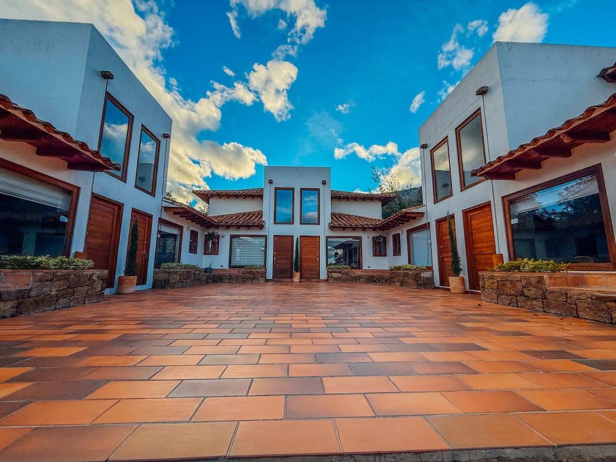 A charming outdoor courtyard is framed by two buildings featuring wooden doors and large windows. A tiled pathway leads towards the entrance, under a sky dotted with clouds, while planters enhance the natural setting.