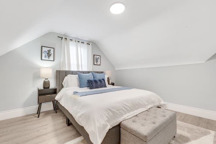 Მყუდრო ორადგილიანი საწოლი პლუშის ბალიშებით, რბილი გდებით, ტუფტის სკამით, საწოლისპირა დახვეწილი მაგიდით, თანამედროვე ლამპით, კედლის ჩარჩოში ჩასმული ხელოვნებითა და გამჭვირვალე ფარდებით ნათელ სხვენის ოთახში.