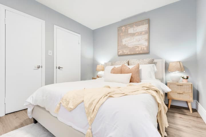 Მოიწვიეთ საწოლი პლუშის ბალიშებით, მყუდრო გდება, საწოლთან მდგარი მაგიდები, ელეგანტური ლამპები, რბილი ხალიჩა და გემოვნებიანი კედლის ხელოვნება ნათელ, ფართო ოთახში.