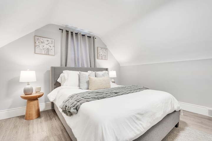 Ფართო საწოლი პლუშის ბალიშებით, მყუდრო ნაქსოვი გდებით, საწოლისპირა დახვეწილი მაგიდებით, თანამედროვე ლამპებით, რბილი ფარდებითა და კედლის ელეგანტური ხელოვნებით მყუდრო სხვენის ოთახში.