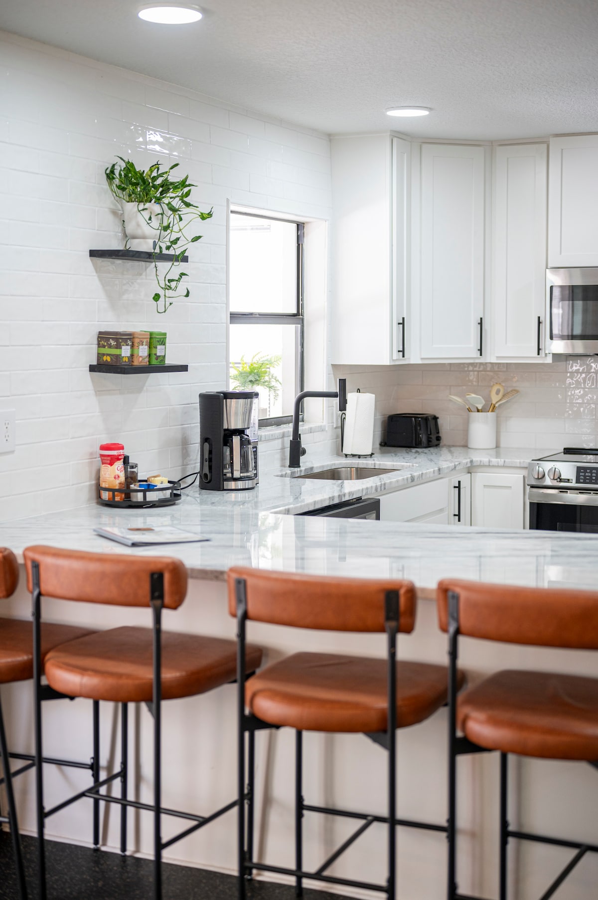 A modern kitchen is featured with sleek white cabinetry and a polished marble countertop. A small shelf on the wall holds potted plants and kitchen essentials. Four barstools with brown seats are arranged at the counter, providing a casual dining option.