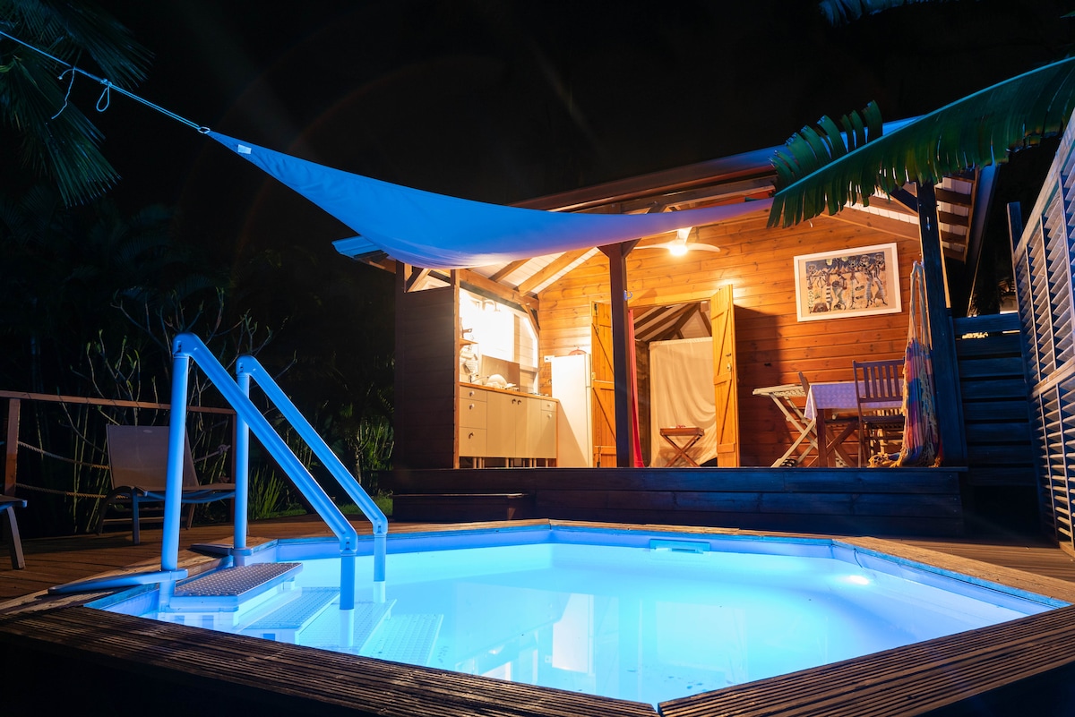 A private pool is illuminated in soft blue light, surrounded by wooden decking. A charming bungalow is visible in the background, featuring a well-equipped kitchen and seating area. Lush greenery frames the area, creating a serene outdoor space, perfect for relaxation.