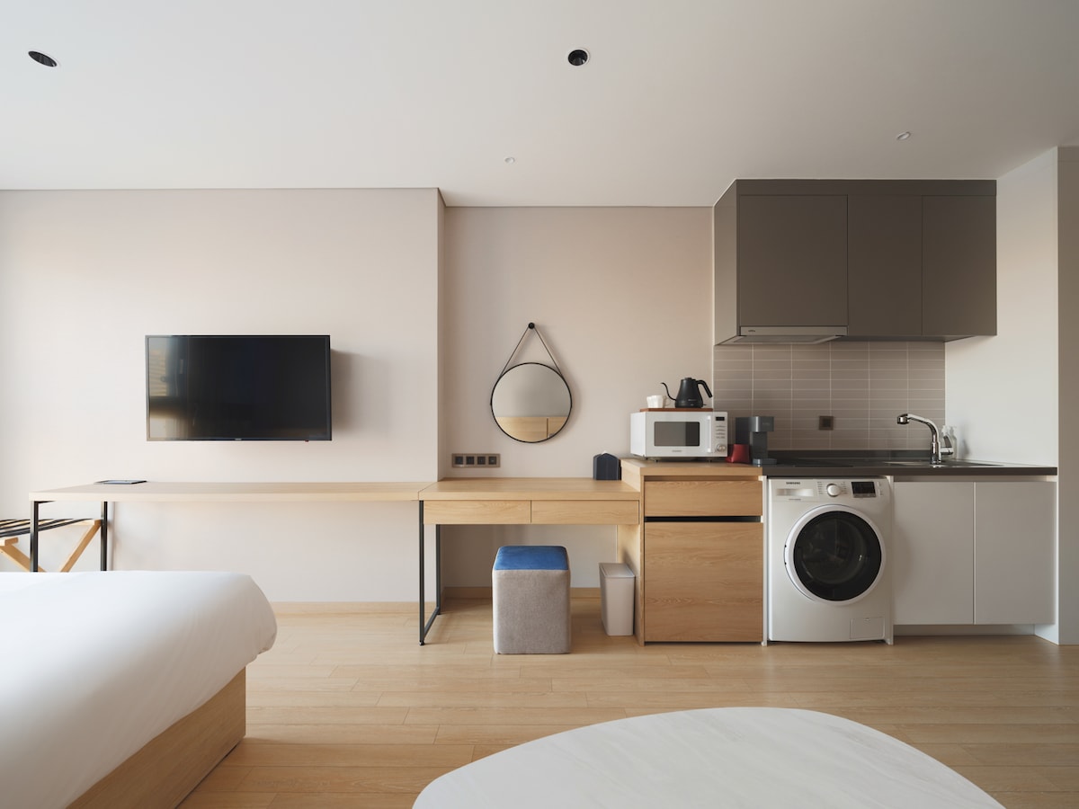 A modern studio space is displayed, featuring a well-equipped kitchenette with a microwave and a washing machine. A neatly arranged wooden table is positioned beneath a wall-mounted television. A round mirror reflects natural light, enhancing the room's functionality and design.