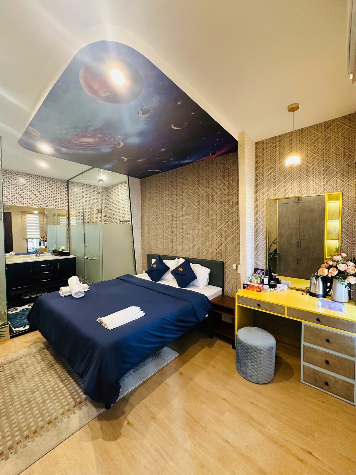 A modern bedroom features a spacious bed with deep blue bedding, accented by two decorative pillows. A mirrored wardrobe and vanity with a yellow finish are visible, along with a floral arrangement. A starry ceiling adds an intriguing touch to the inviting atmosphere.