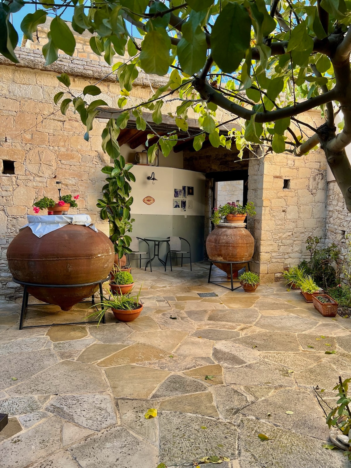 An entrance area is framed by rustic stone walls, featuring two large clay vessels placed on metal stands. Potted plants decorate the space, and a small table with chairs is visible beyond the entrance, creating an inviting outdoor atmosphere.