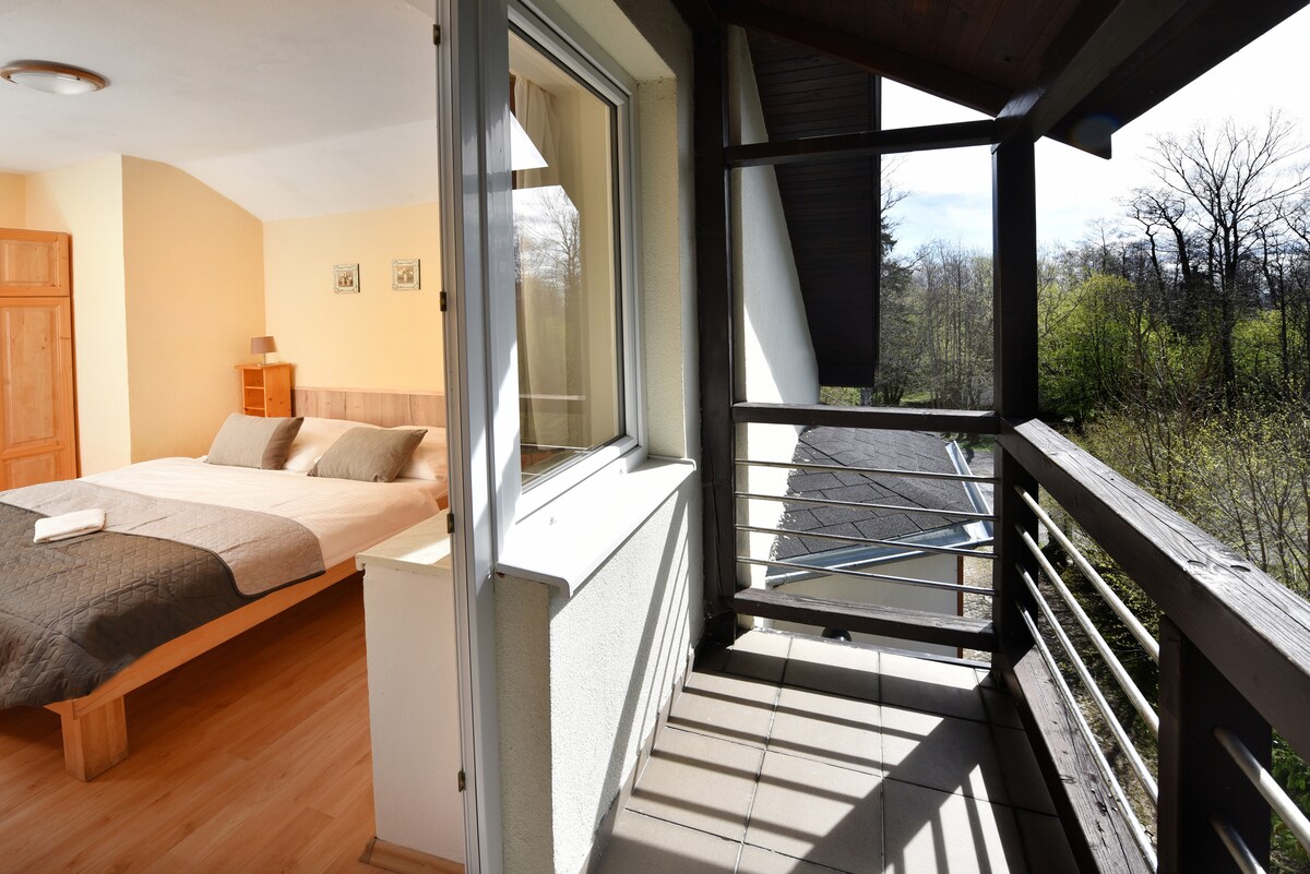 A spacious bedroom is visible through a large window, featuring a cozy bed dressed in neutral tones. Adjacent, a private balcony offers views of surrounding greenery, with sunlit tiles creating an inviting outdoor space. Natural light filters through, enhancing the serene ambiance.