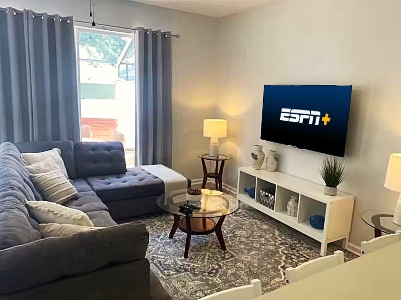 A comfortable living room is presented with a spacious sectional sofa in gray, accompanied by a round glass table. Soft curtains frame the window, allowing natural light to brighten the space. A television is mounted on the wall, offering access to streaming services.