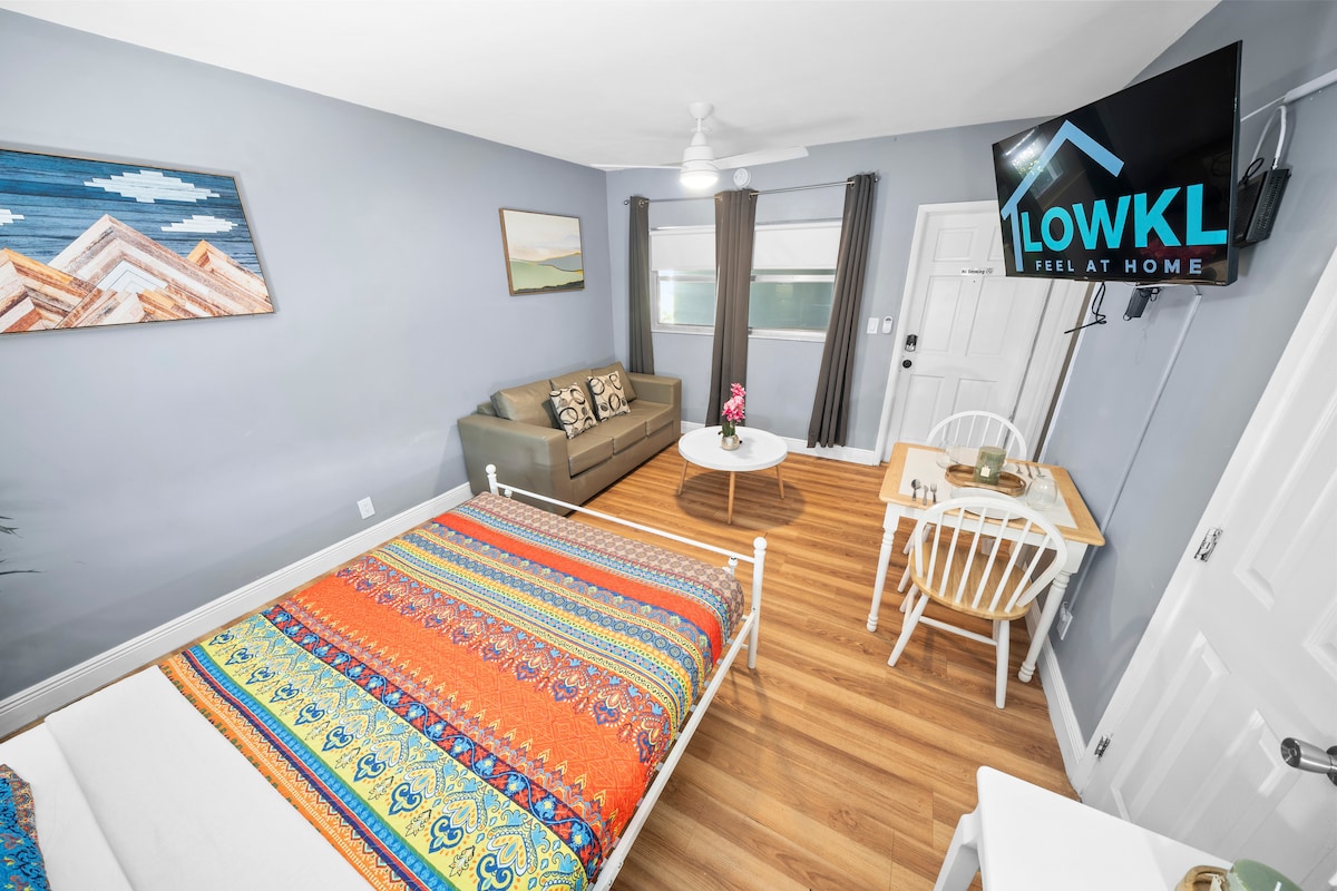 A well-lit studio space is presented with a vibrant, patterned bedspread covering a spacious bed. A comfortable sofa and coffee table are positioned nearby, complemented by a dining table with two chairs. A flat-screen TV is mounted on the wall, while large windows provide natural light.