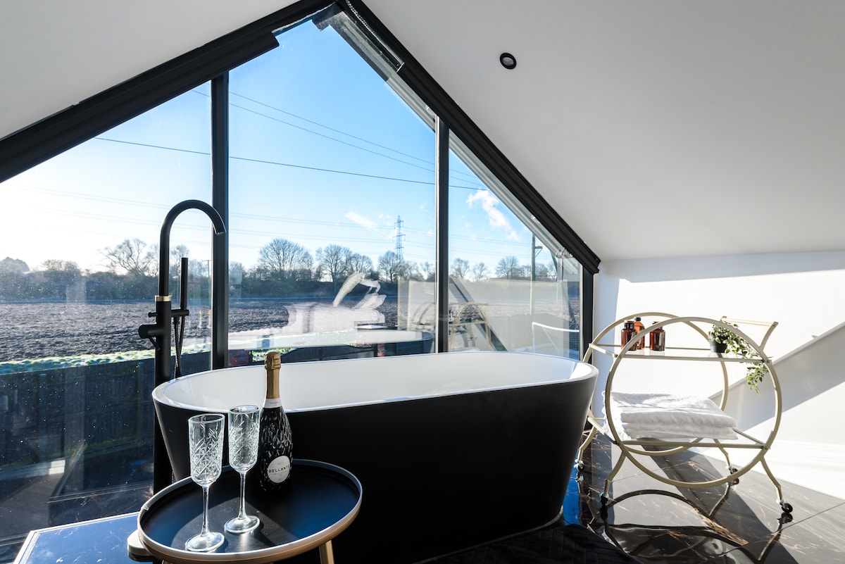 A freestanding bath is positioned near large windows, offering views of the surrounding farmland. A small table holds two champagne flutes and a bottle, while a cozy chair is placed softly against the wall, enhancing the tranquil setting of the space.