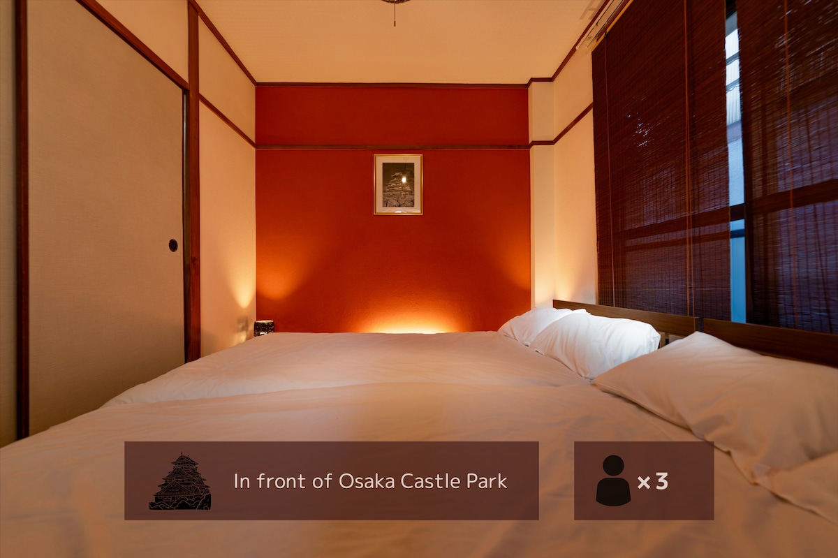 A warm-toned bedroom is presented, featuring a spacious bed with crisp white linens. The walls are a calming shade of red, complemented by decorative accents. Natural light filters in through bamboo shades, creating a serene ambiance for relaxation.