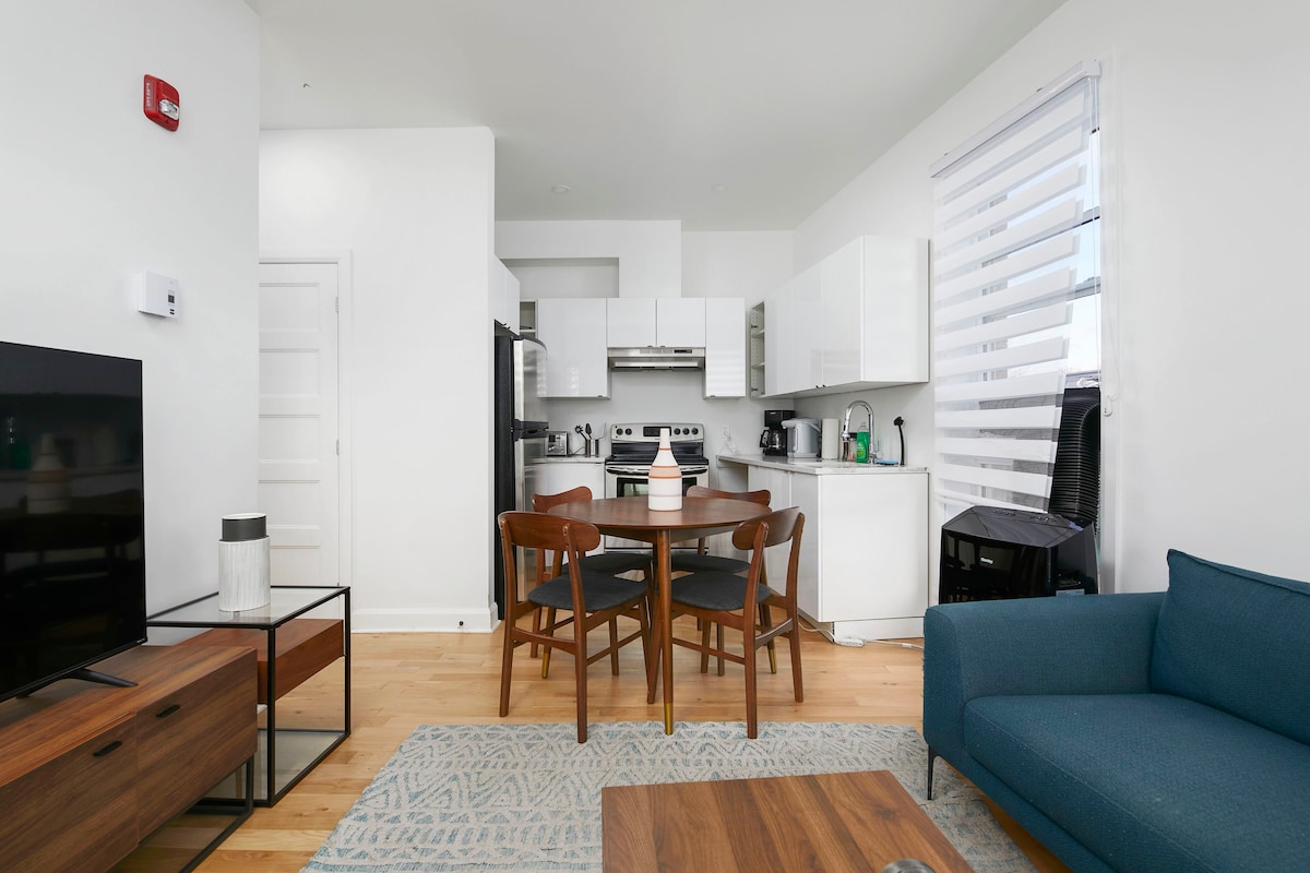 An open living area features a modern dining table with four wooden chairs, a stylish blue couch, and a wooden coffee table. A fully equipped kitchen with white cabinetry and stainless-steel appliances is visible in the background, illuminated by natural light from the window.