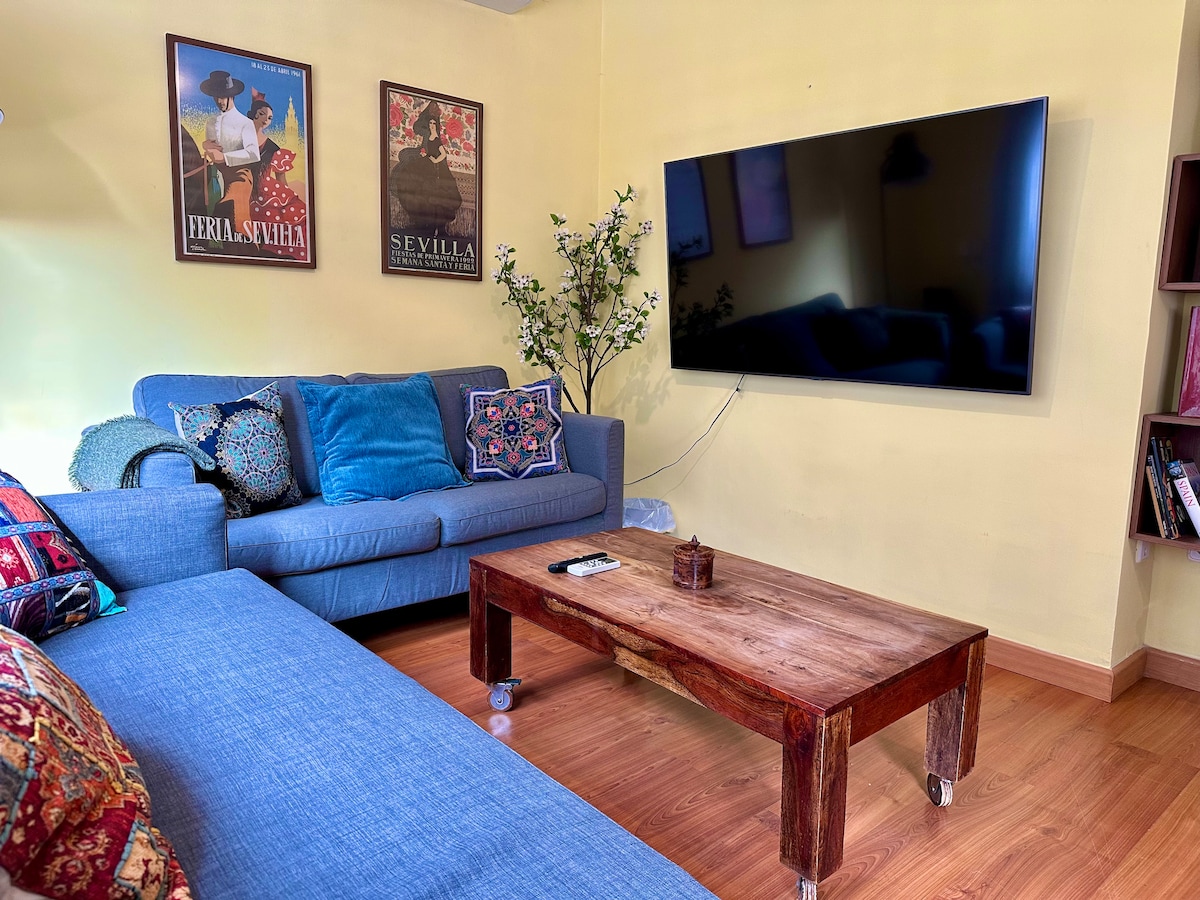 The living area features a comfortable gray sofa adorned with colorful cushions, complemented by a rustic wooden coffee table. A large television is mounted on the wall, and the space is brightened by warm yellow walls, creating a cozy environment for relaxation.