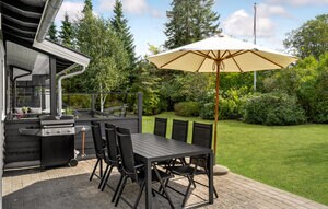 An outdoor dining area is situated on a patio, featuring a large table with eight chairs beneath a shaded umbrella. A barbecue grill is positioned nearby, and a well-maintained garden surrounds the space, offering a tranquil view of greenery and trees.