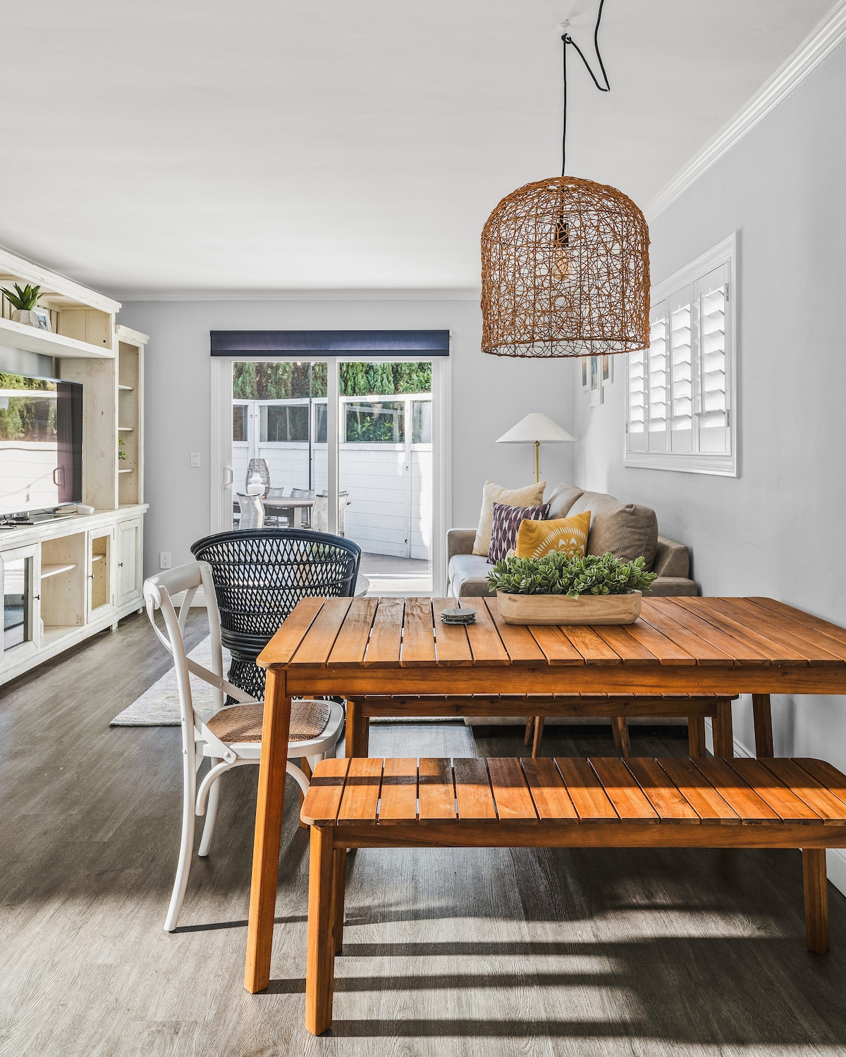 A wooden dining table is paired with a bench and a white chair, offering seating for casual meals. A woven pendant light hangs overhead, illuminating the space. A comfortable sofa can be seen in the background, creating a relaxed living area that invites leisure.