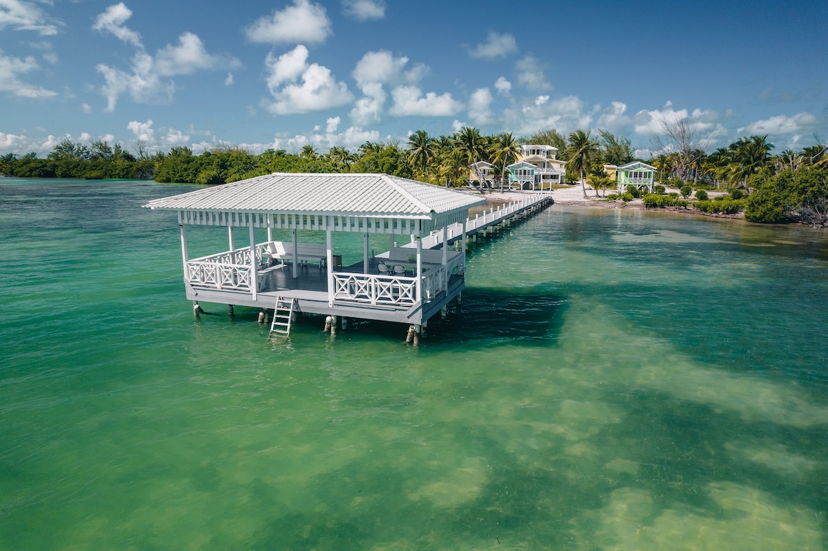 A wooden deck extends over clear turquoise waters, featuring a covered gazebo with multiple seating areas. Lush greenery and palm trees can be seen in the background, creating a serene coastal environment. A sandy shore is visible on the right side of the image.