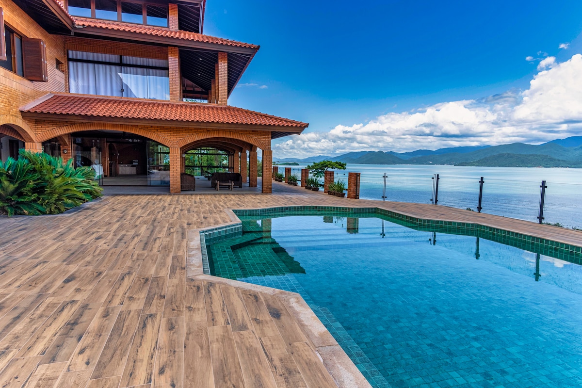 An outdoor pool area is presented with clear water reflecting the blue sky. Surrounding the pool, wooden decking leads toward a brick structure, showcasing large windows and a terrace. Lush vegetation is visible in the background, enhancing the natural scenery.