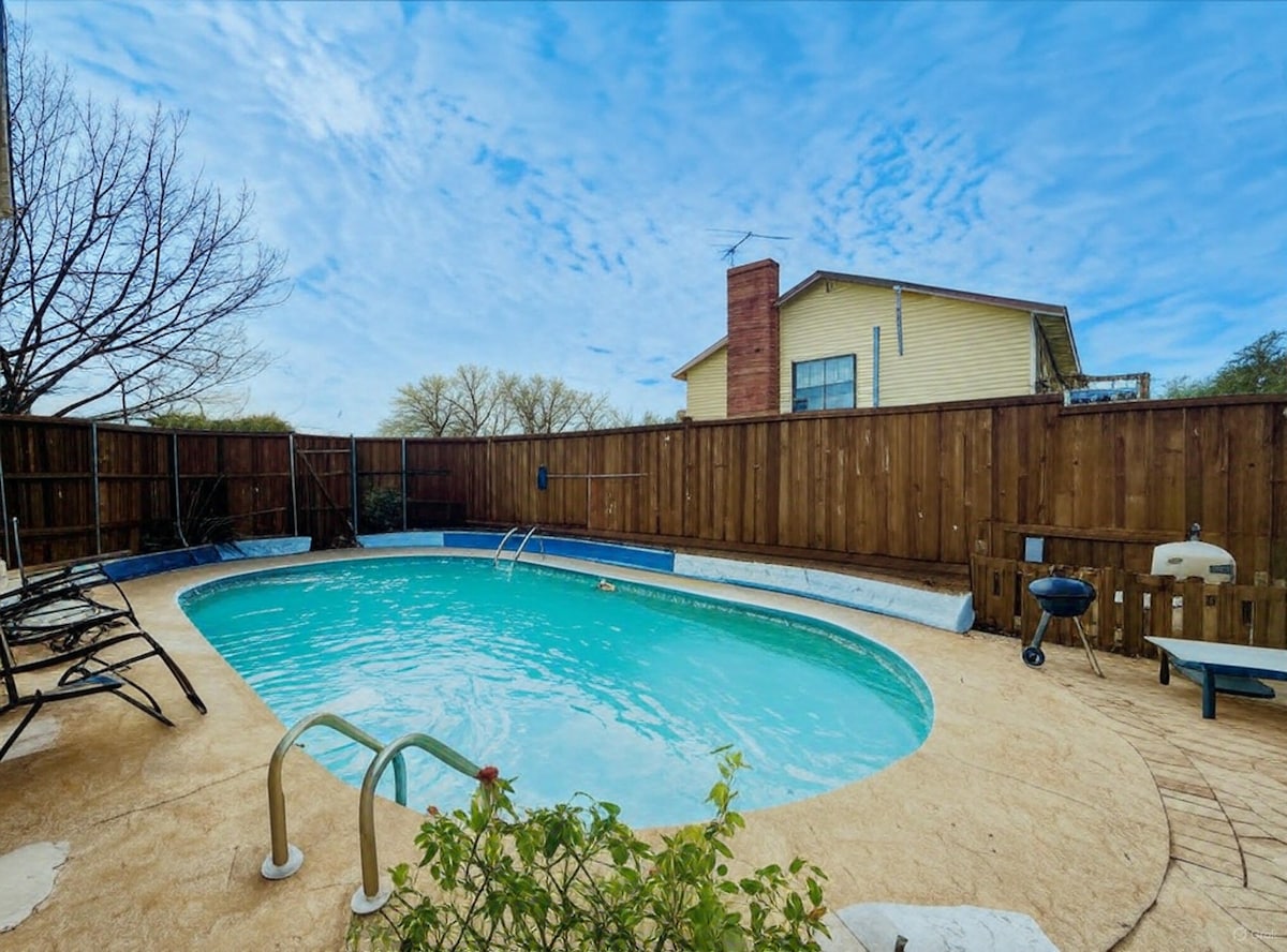 A serene backyard pool area is enclosed by a wooden fence. The clear blue water is complemented by a surrounding tan deck. Lounge chairs are positioned beside the pool, while a grill is situated nearby for outdoor cooking.