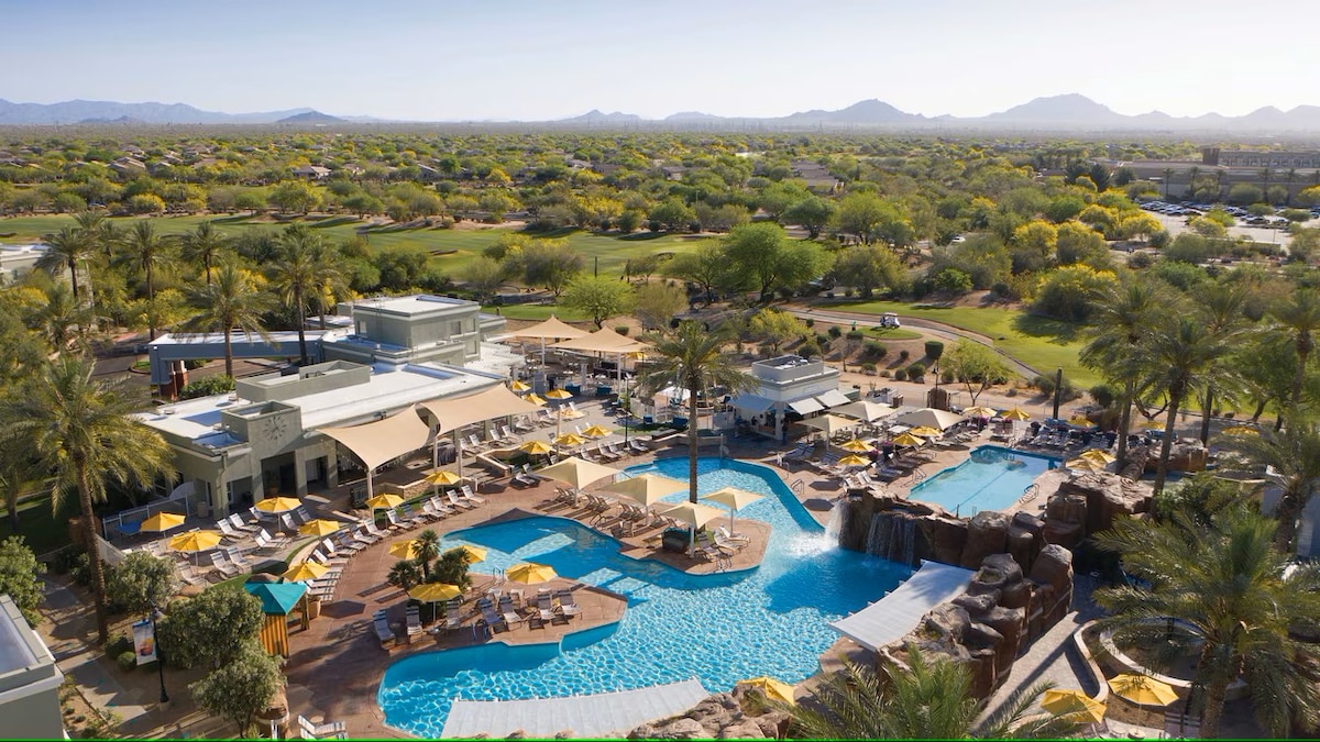 An overview of the resort's expansive pool area is presented, featuring multiple swimming pools, a waterfall feature, and lounge chairs surrounding the water. Lush greenery and palm trees create a tropical ambiance, while the distant mountains provide a scenic backdrop.