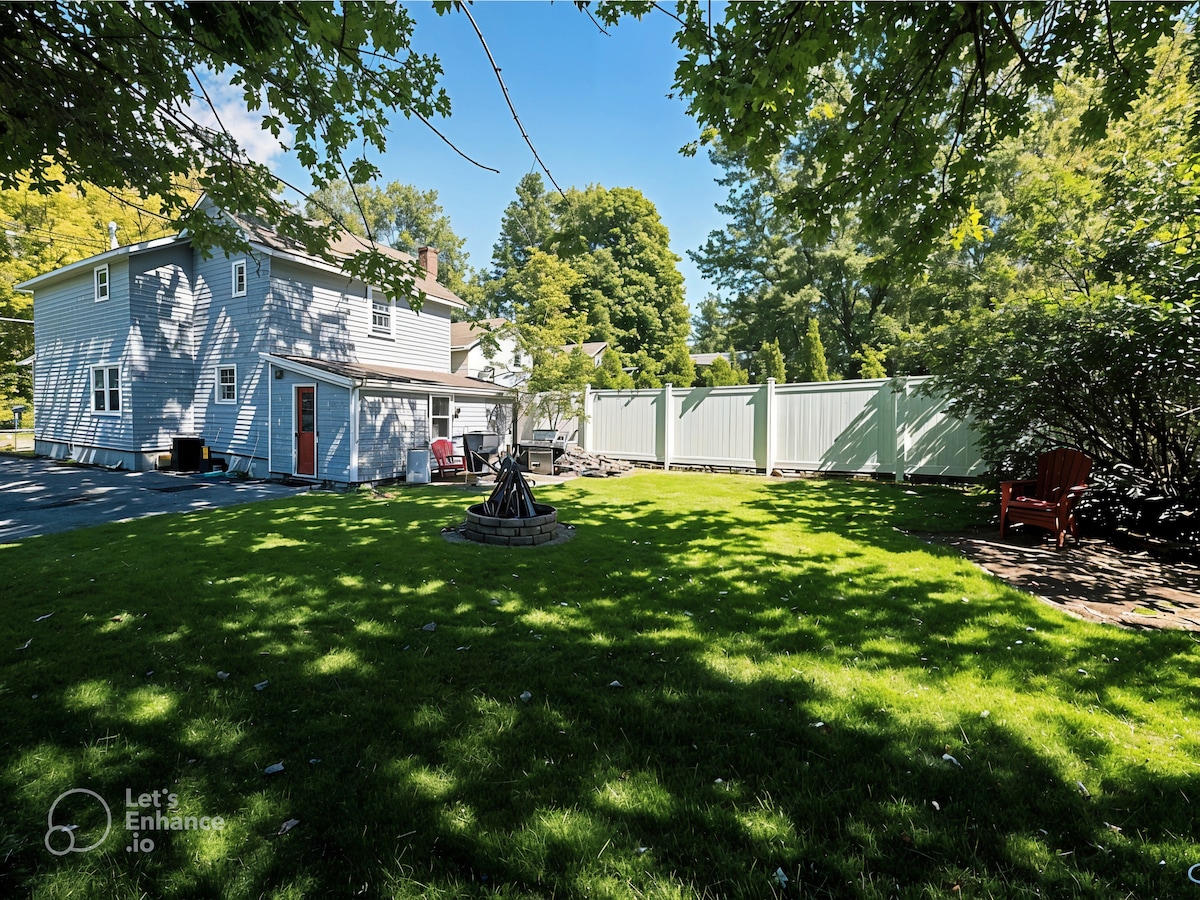 A spacious backyard is featured, showcasing a well-manicured lawn surrounded by leafy trees. A fire pit is positioned at the center, with seating around it. Fenced areas provide privacy, and a cozy red chair is visible under the shade, enhancing the outdoor setting.