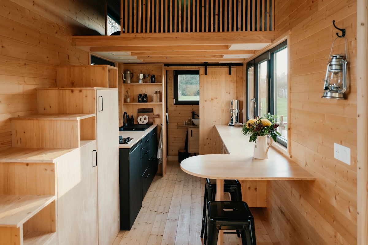 The interior of the tiny house showcases a warm wood-paneled space, featuring a compact kitchen equipped with essential appliances. A small dining area with a round table and black stools faces large windows, allowing natural light to fill the room. A staircase leads to the mezzanine bedroom.