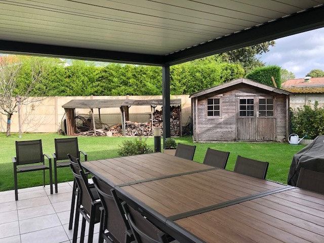 A covered terrace features a large wooden dining table surrounded by several chairs. In the background, a well-maintained garden is visible, bringing attention to a wooden shed and a storage area with neatly stacked firewood.