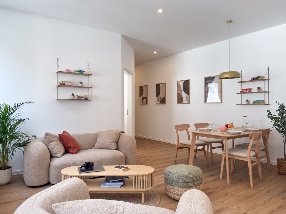 An inviting living space features soft, rounded sofas arranged for comfort. A light wood dining table with modern chairs is set for a meal. Wall shelves display decorative items, while a potted plant adds a touch of greenery to the room.