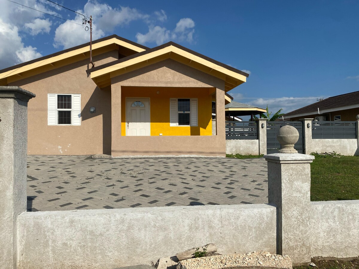 The exterior of the house features a modern design with a light brown stucco finish and a bright yellow accent wall. A spacious driveway is paved with geometric patterns, bordered by a low stone wall with a round pillar at the entrance.