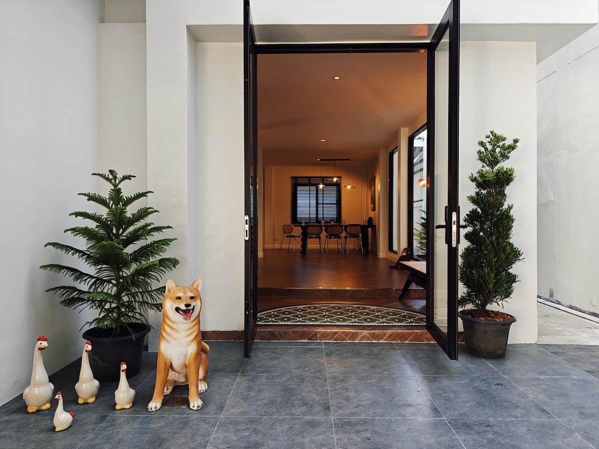 The entrance features a pair of large glass doors that open into a spacious interior. A decorative dog statue and garden plants are positioned outside, enhancing the welcoming ambiance. The flooring is tiled, leading to a wooden dining area visible through the doorway.