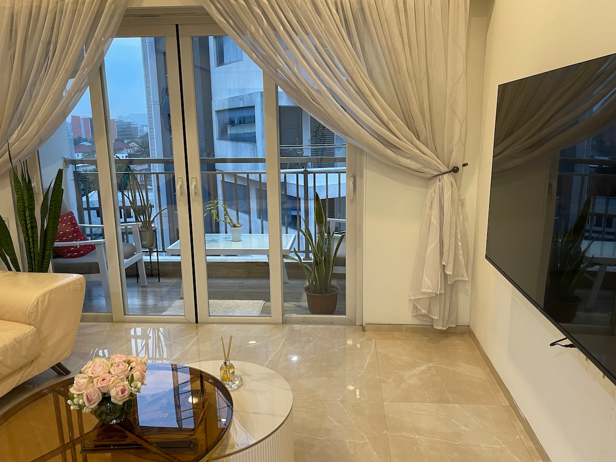 A modern living room is presented, featuring a light-colored sofa and a stylish glass coffee table. Large sliding glass doors open to a balcony, allowing natural light to pour in. Sheer curtains frame the doors, and potted plants add a touch of greenery.