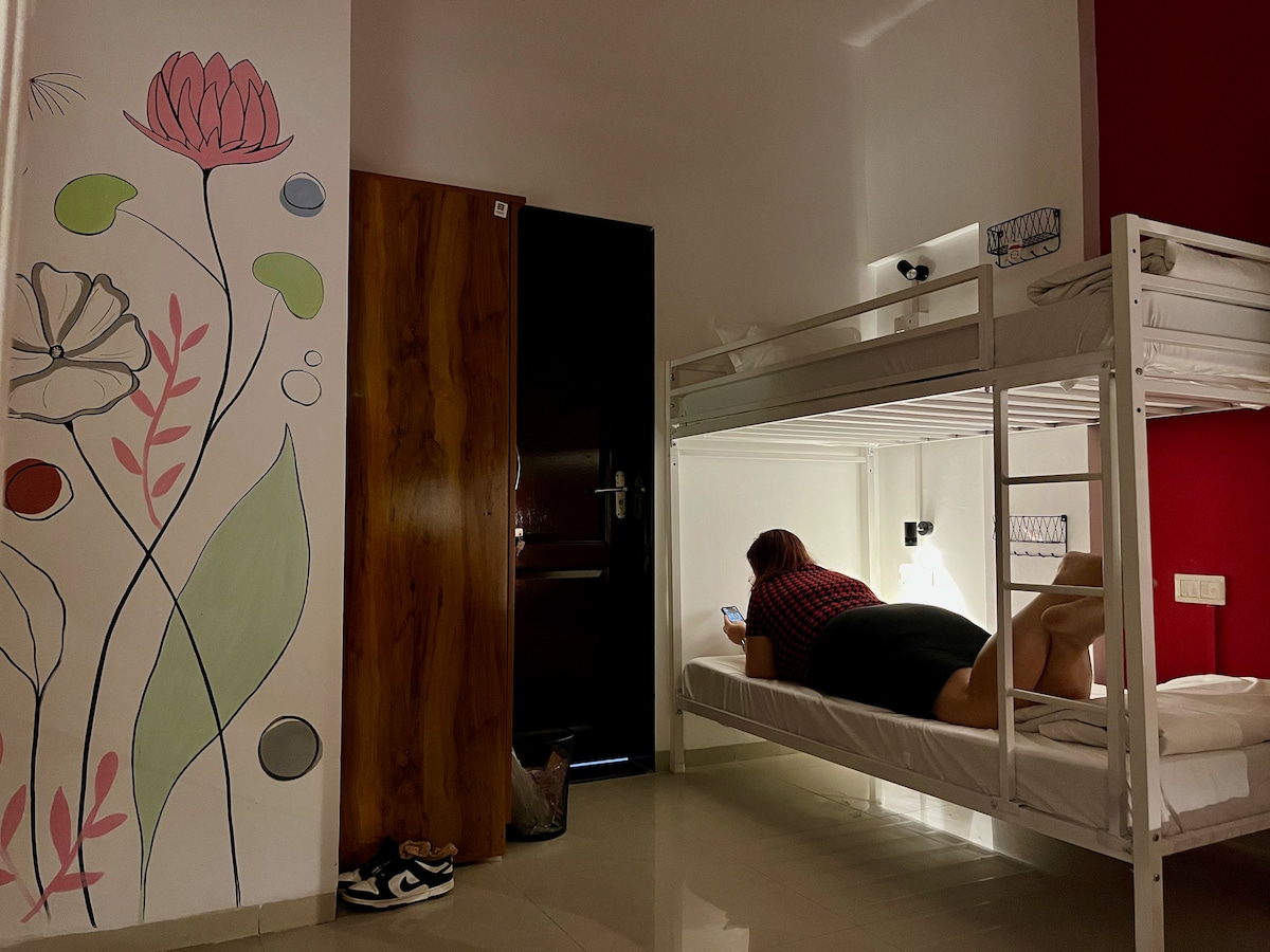 A spacious room features a bunk bed against a wall adorned with hand-painted floral designs. Soft lighting illuminates the area, while a guest is seen reclining on the lower bunk, engaged with a mobile device. A dark wooden door and subtle floor tiles complete the setting.