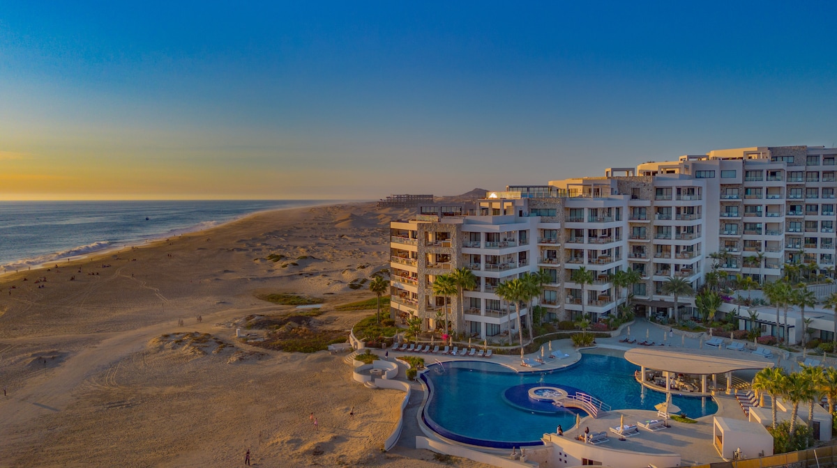 A beachfront resort complex is depicted, featuring modern architecture and expansive pool areas. The setting includes sandy shores and the ocean, shown under a gradient sky as the sun sets. Lush palm trees surround the pool, creating a tropical ambiance.