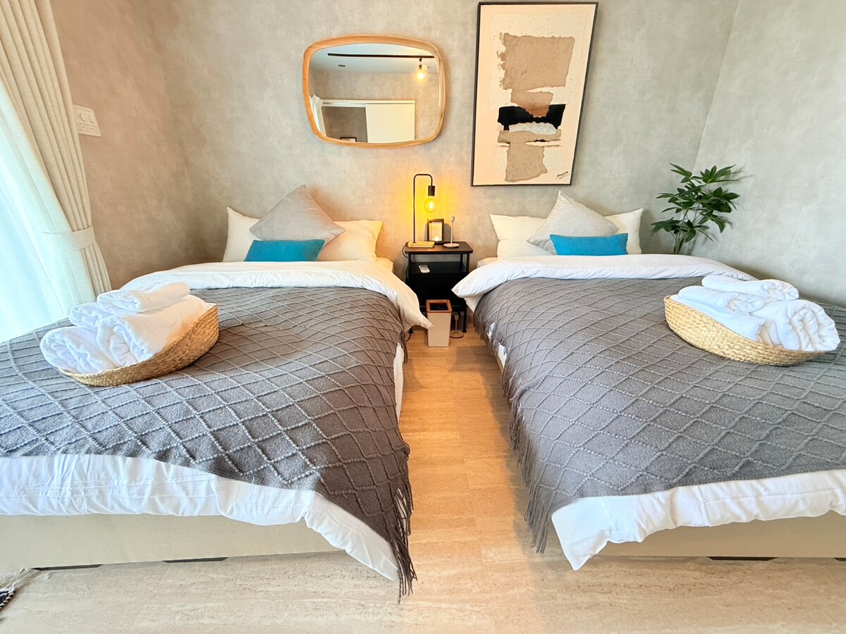 A well-appointed guestroom features two neatly arranged beds, each topped with crisp white linens and textured gray throws. A small nightstand with a lamp is positioned between the beds. A stylish mirror and a potted plant add a touch of decor, while natural light flows in through the window.