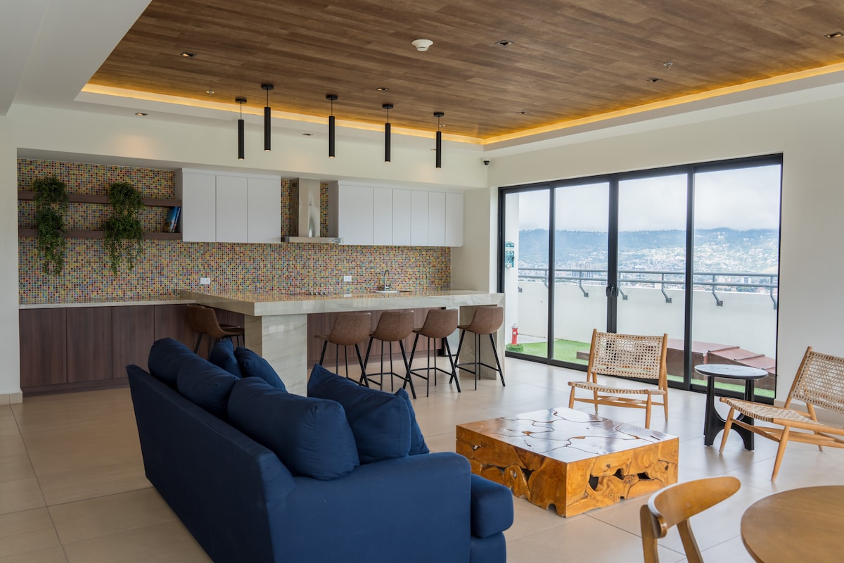 A modern communal area is featured with a spacious kitchen and bar seating. Natural light floods through large windows, highlighting a cozy blue sofa and a wooden coffee table. The vibrant backsplash adds a touch of color to the sleek design while greenery accents the space.